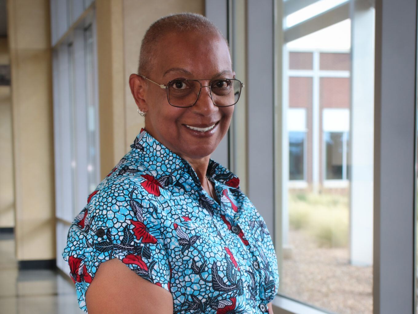 Dee-Dee smiling, wearing glasses and a colorful dress, standing indoors near windows.