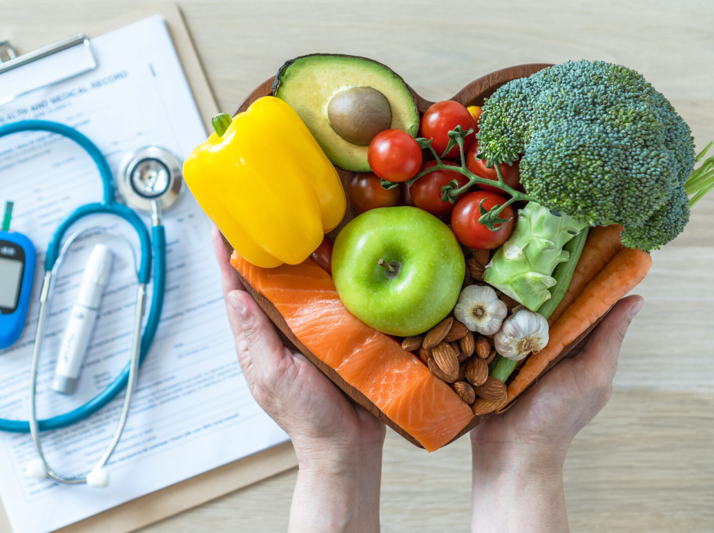 Hands holding a heart-shaped bowl with vegetables and fruits, next to medical equipment and health record clipboard.