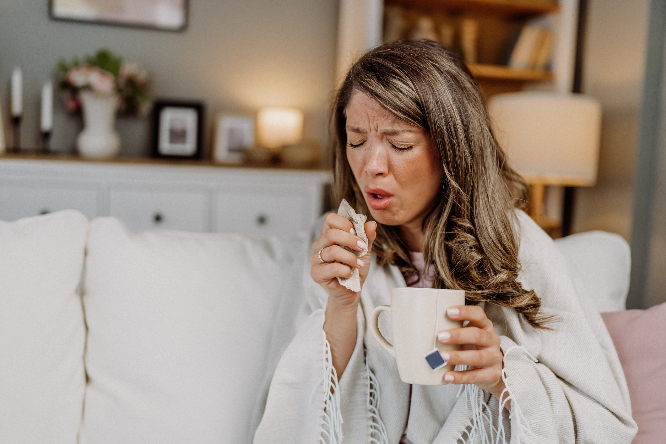 Woman sneezing while holding a tissue and a mug, sitting on a couch.
