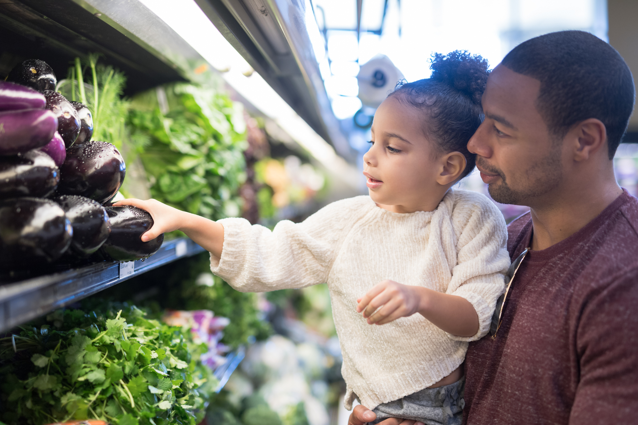 Father holding daughter as she reaches for eggplant in supermarket