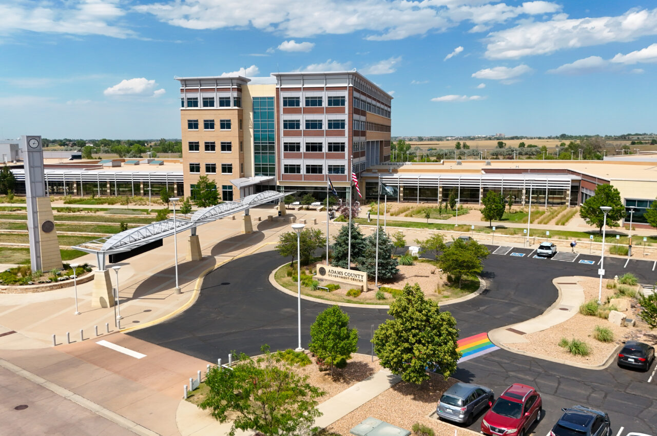 Adams County Government Center with clock tower, rainbow crosswalk, and parking lot under blue sky.