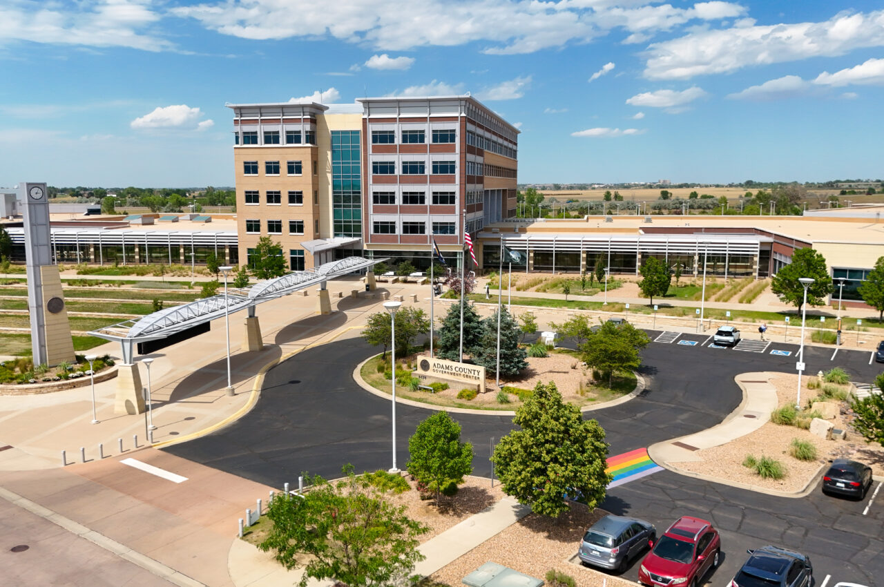 Adams County Government Center with clock tower, rainbow crosswalk, and parking lot under blue sky.