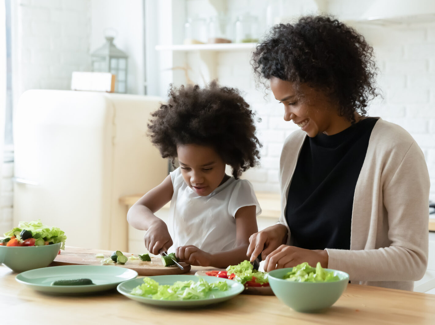 Photo of a mother and child smiling while chopping vegetables together in a bright kitchen.