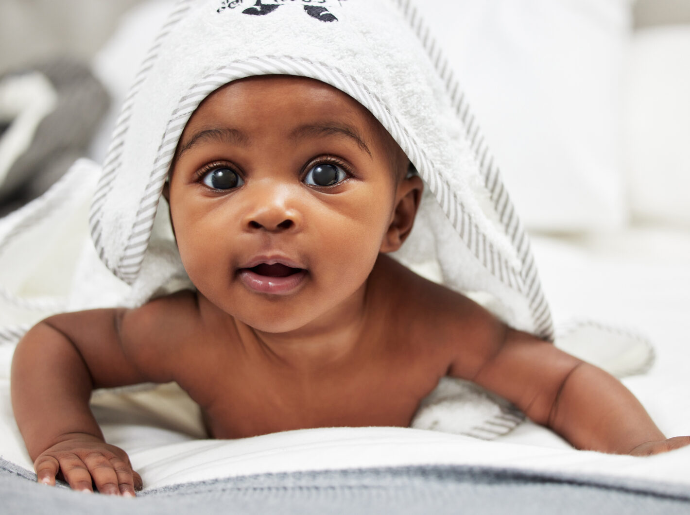 Photo of a baby laying on stomach under white towel, looking forward.