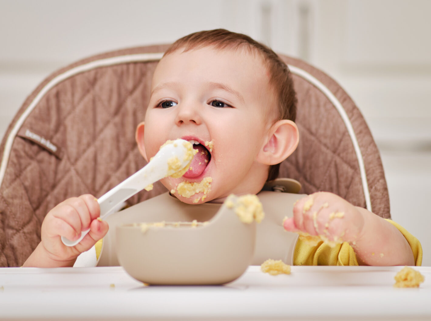 Photo of a baby in high chair eating with spoon, looking delighted, food everywhere.