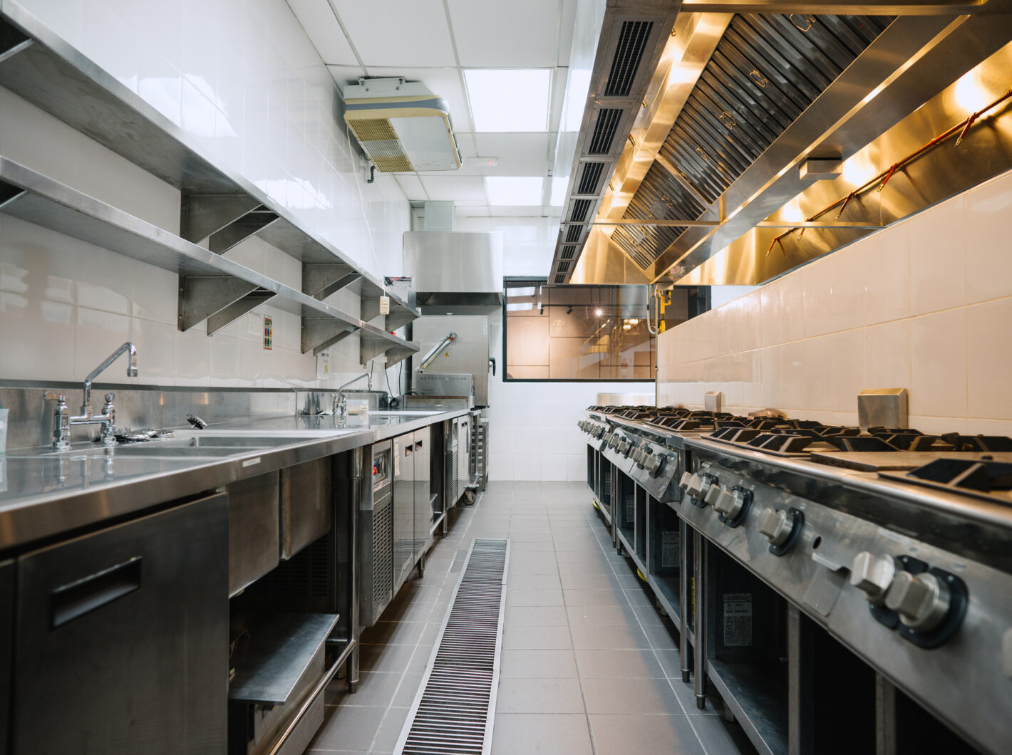 Commercial kitchen with stainless steel appliances, long countertops, overhead ventilation, and industrial ovens on the right.