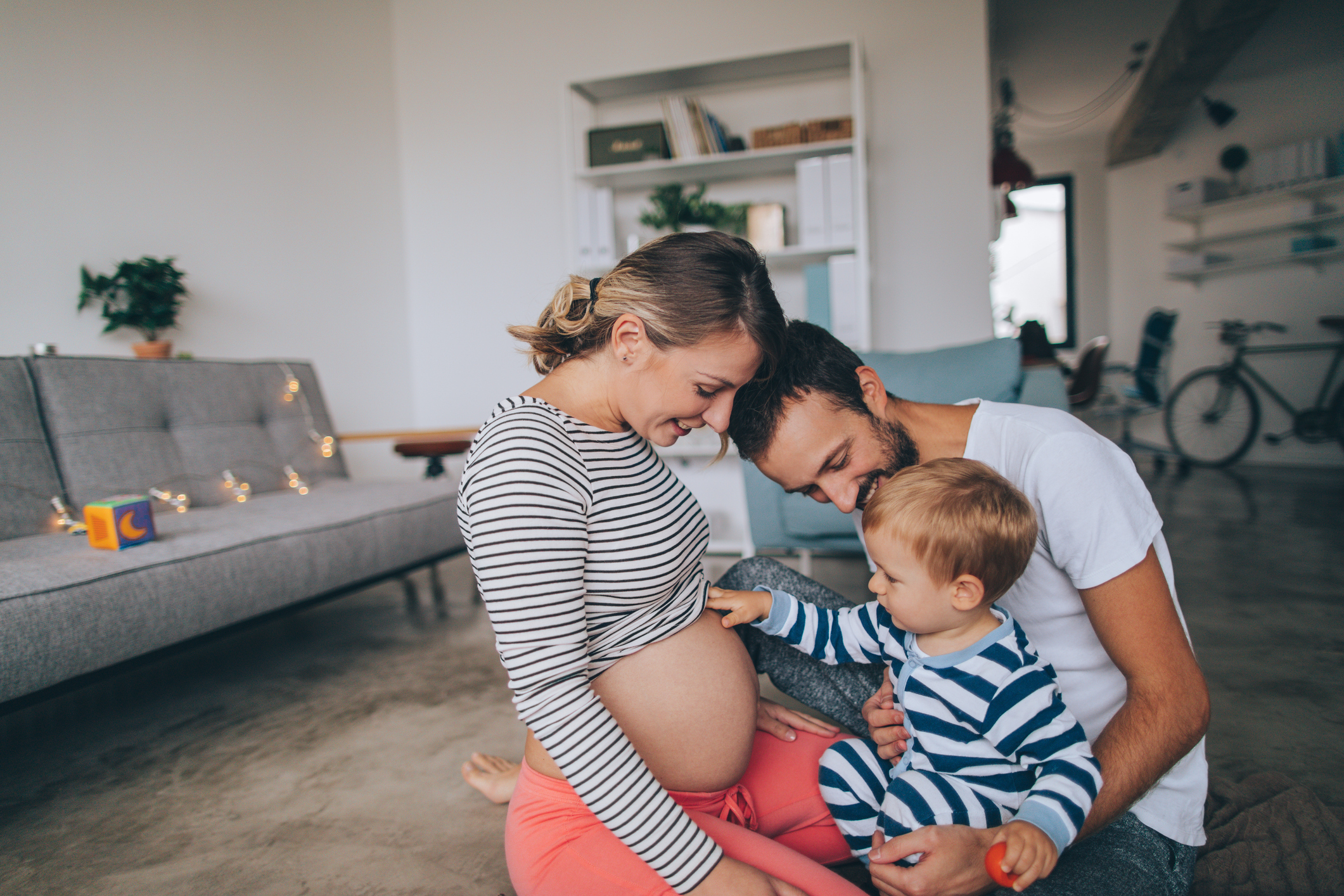 Photo of a pregnant woman with a toddler touching her belly and a smiling man in a cozy living room.
