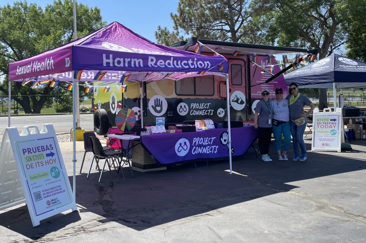 Project Connects booth with "Harm Reduction" and "Sexual Health" banners, offering free STI testing, staffed by three people.
